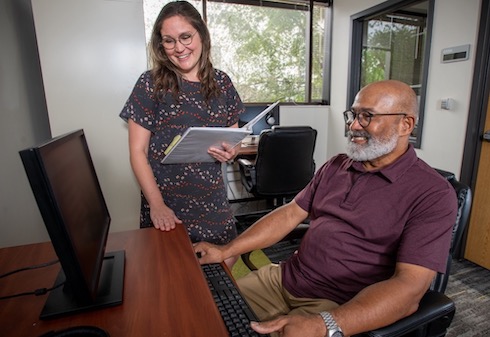 smiling woman holding notebook helps senior man at computer