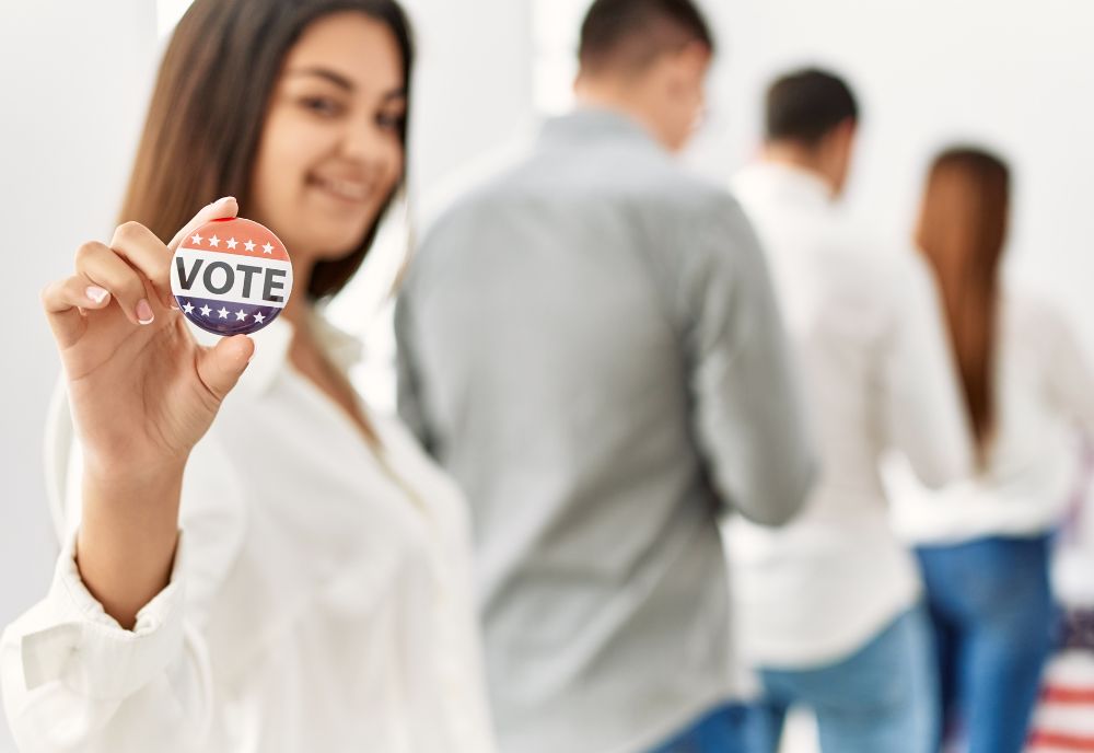 woman holds up Vote button while standing in line. Photo from Adobe Stock