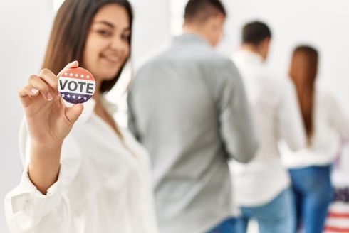 woman holds up Vote button while standing in line. Photo from Adobe Stock