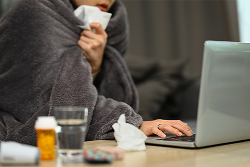 person wrapped in blanket holding tissue and working on laptop