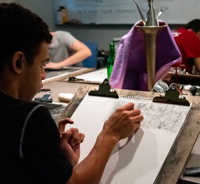 A student is shown drawing with charcoal.