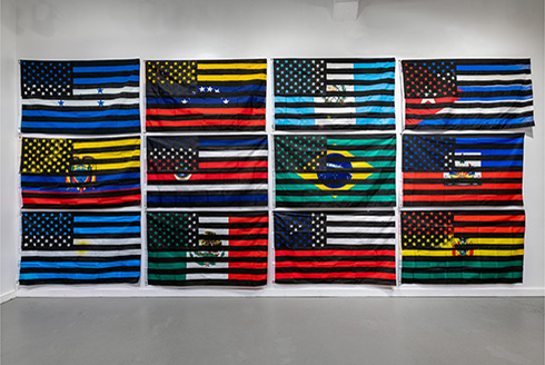 A wall of 12 national flags with black and white American flags screen printed on top of them, by artist Eddy A. López (b. 1978, Matagalpa, Nicaragua), In-Dependencia, 2021-2022. Courtesy of the artist.   