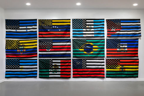 A wall of 12 national flags with black and white American flags screen printed on top of them, by artist Eddy A. López (b. 1978, Matagalpa, Nicaragua), In-Dependencia, 2021-2022. Courtesy of the artist.   