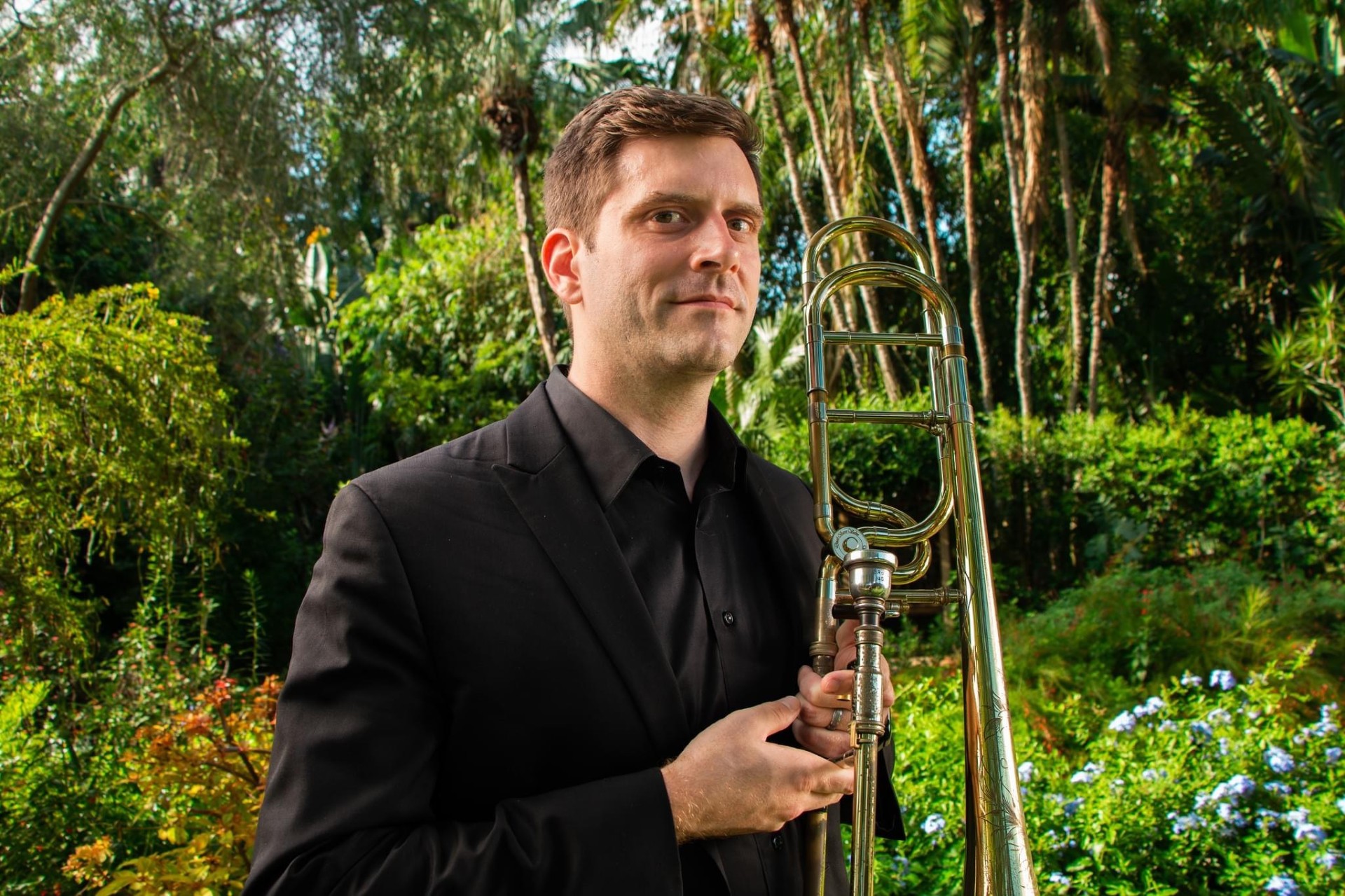 Headshot image of Ross Holcombe holding a trombone and standing in front of natural foliage.