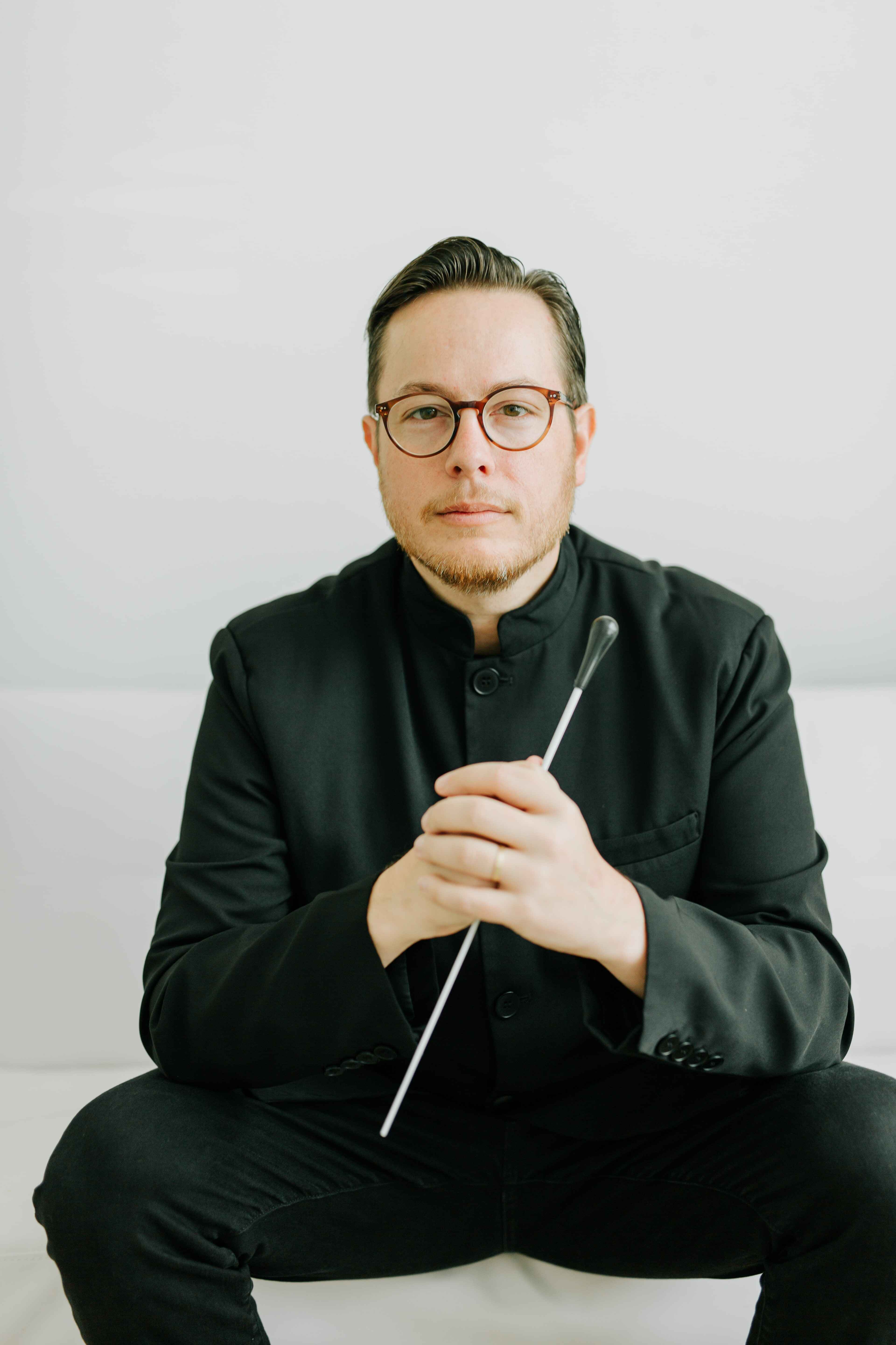 Matthew Wardell sitting on a chair and holding a conducting baton.