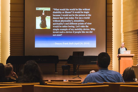 A person is shown speaking at a lectern in a hall while slides are shown.