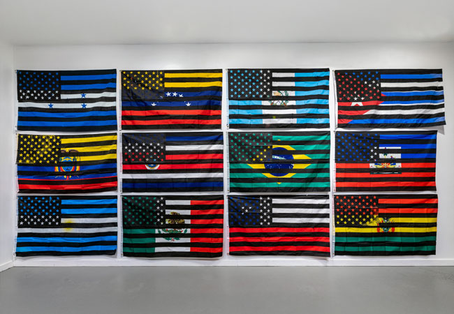 A wall of 12 national flags with black and white American flags screen printed on top of them, by artist Eddy A. López (b. 1978, Matagalpa, Nicaragua), In-Dependencia, 2021-2022. Courtesy of the artist.   