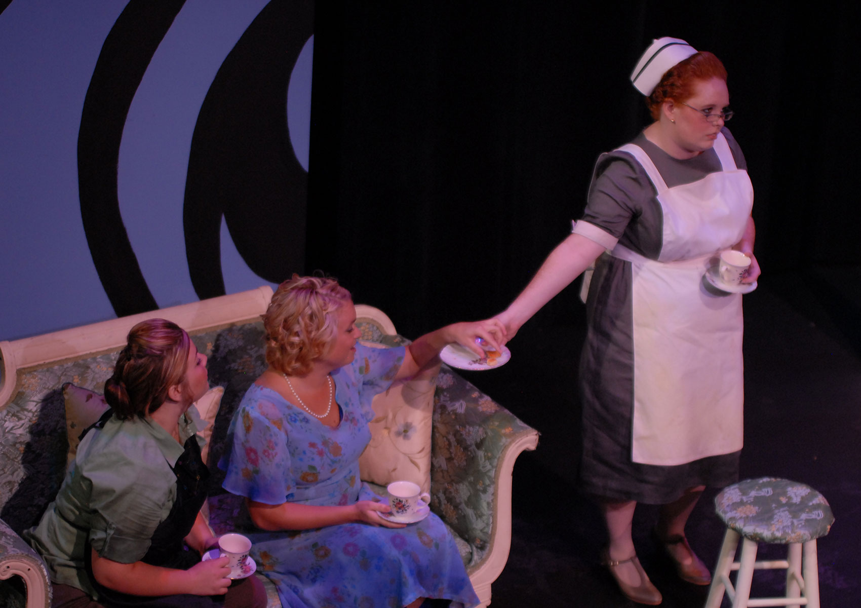 Two women sit on a couch while the woman on the right accepts a cookie from a saucer held by a maid, who looks away from the woman toward the audience while holding a teacup and saucer.