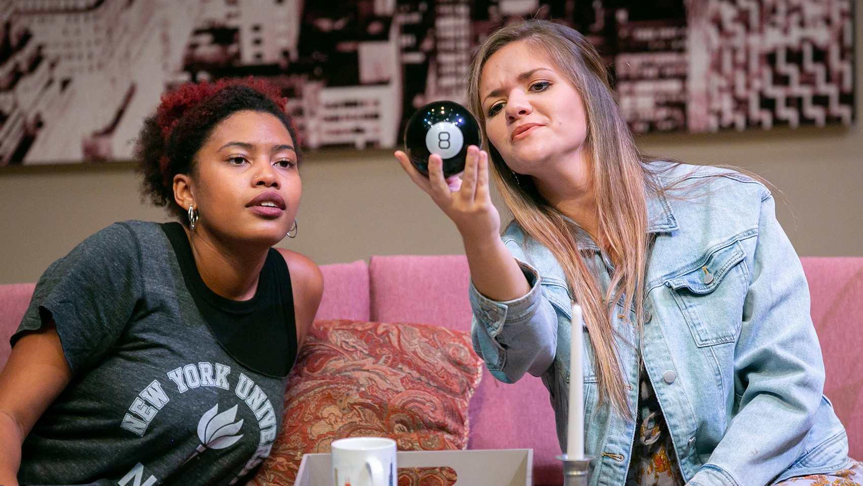 The two main women characters sit on a couch observing a magic eight ball.
