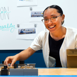 The image shows a smiling person with glasses and a light-colored cardigan working on an architectural model.