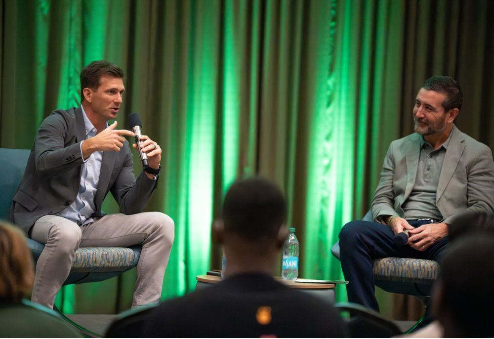 Pete Petersen speaks into a microphone sitting on stage next to Allen Clary