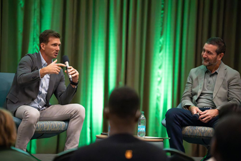 Pete Petersen speaks into a microphone sitting on stage next to Allen Clary