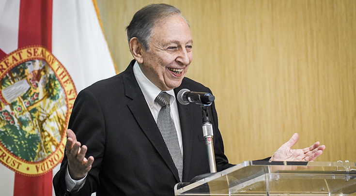 Robert Gallo, MD, stands at a lecturn and gives a speech with the Florida flag in the background.