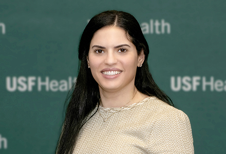 A woman with long dark hair wearing a gold shirt on a green background. 