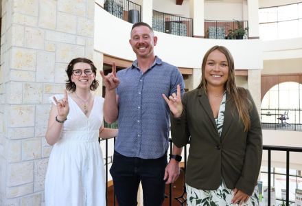 Caroline Merrriman (podcast interviewer), Jordan Howell, and Kaylee Eckelman smile with the "Go Bulls" sign in the Sarasota campus atrium