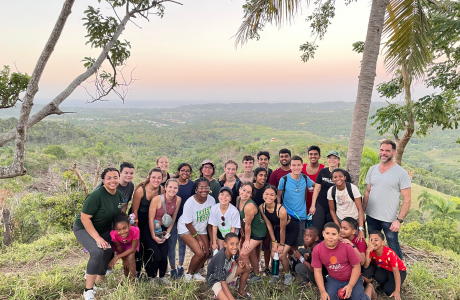 Honors students and faculty smile with children in the Dominican Republic overlooking a picturesque landscape.