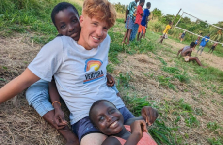 Honors student smiles while sitting in the grass with two children from Ghanna. 