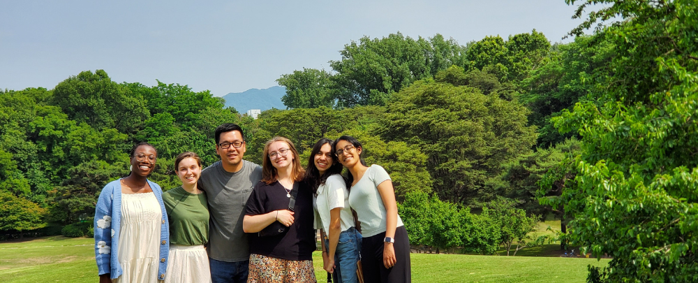 Students and faculty smile in front of lush trees and a mountain range.
