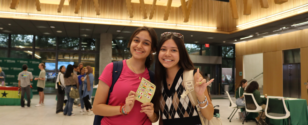 Two honors student smile in the atrium while holding a Tapas nigh information card