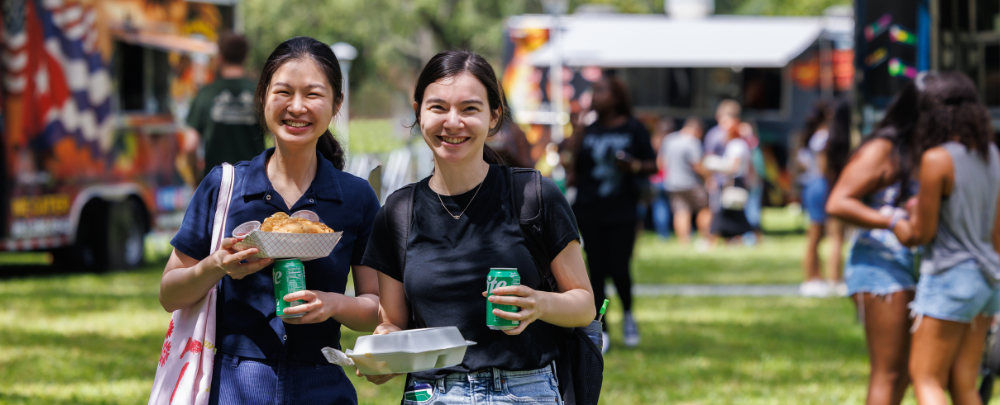 Two students smile while holding sodas and food from food trucks