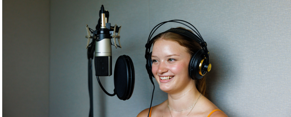 Student smiles wearing headphones and standing in front of a microphone in the multimedia studio.