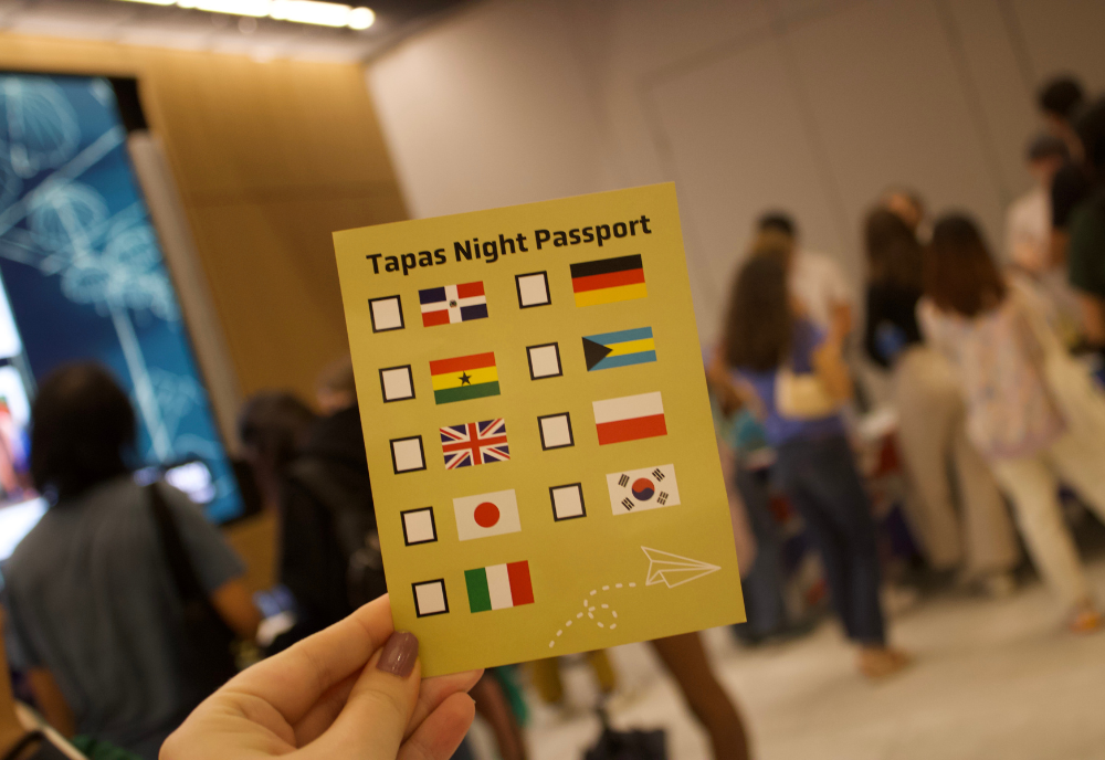 A student holds up a Tapas Night passport card. In the background students visit different booths. 