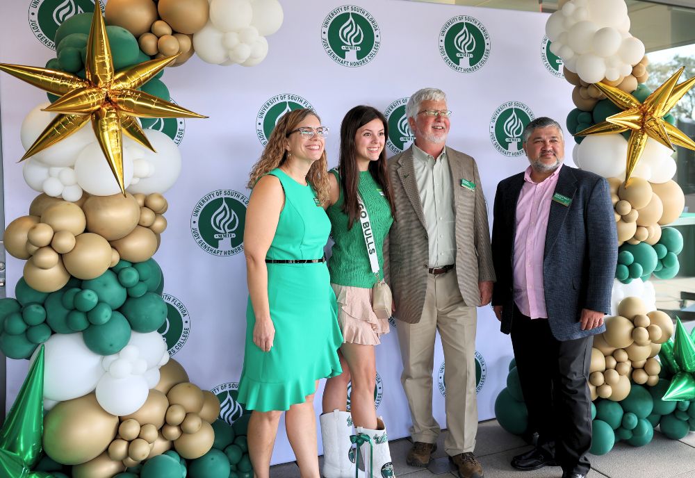 Lindy Davidson, Honors alum Avalon Jade Theisen, Dean Adams, and advisor Arnaldo Mejias pose in front of a backdrop on the patio