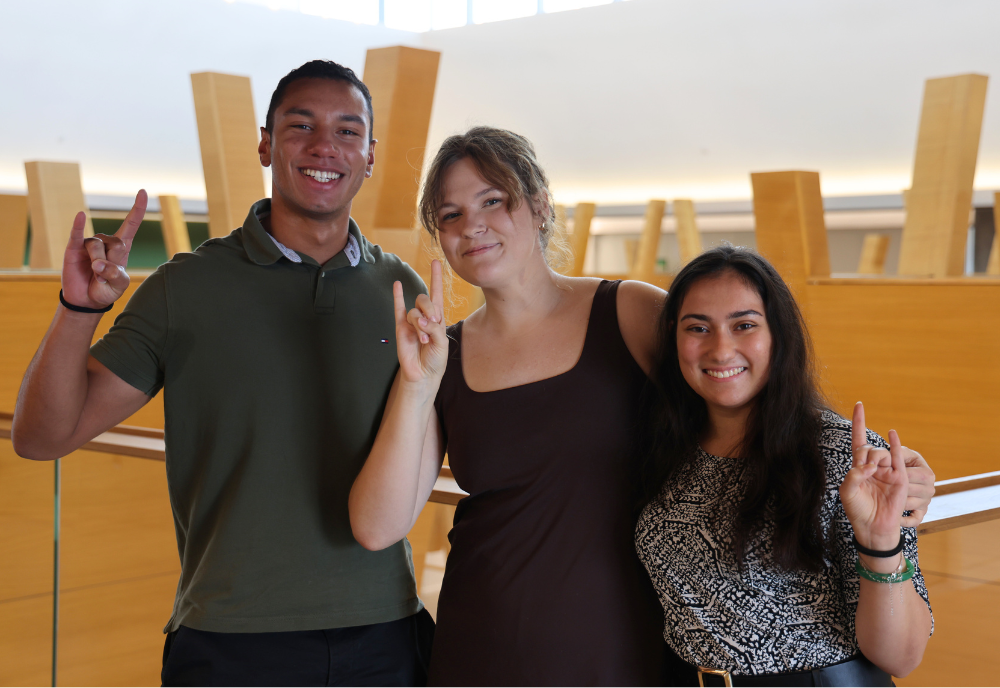 Judy Genshaft Honors College students Breno Nery Dantas, Jamie Powers, and Reva Gandhi pose while making "Go Bulls" hand signs inside of the Honors College building.