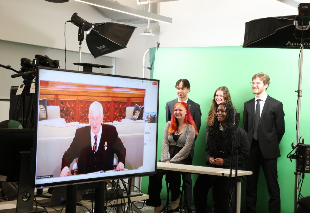 Five Honors students stand in front of a green screen while listening to Right Honorable Sir Lindsay Hoyle from a monitor.