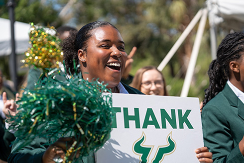 A small group of USF students cheer while holding gold and green poms and a white and green Thank 