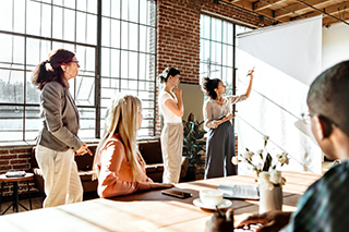  group of people are engaged in a presentation or meeting in a well-lit, modern office with large windows and exposed brick walls.