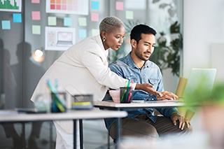 two people sit at a computer in an office setting working on a project