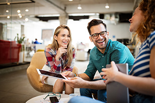 a group of three people in a bright, modern office setting. They are sitting together in a casual arrangement, engaged in a lively and cheerful conversation.