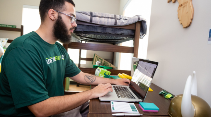 college student sits in dorm room at desk on computer