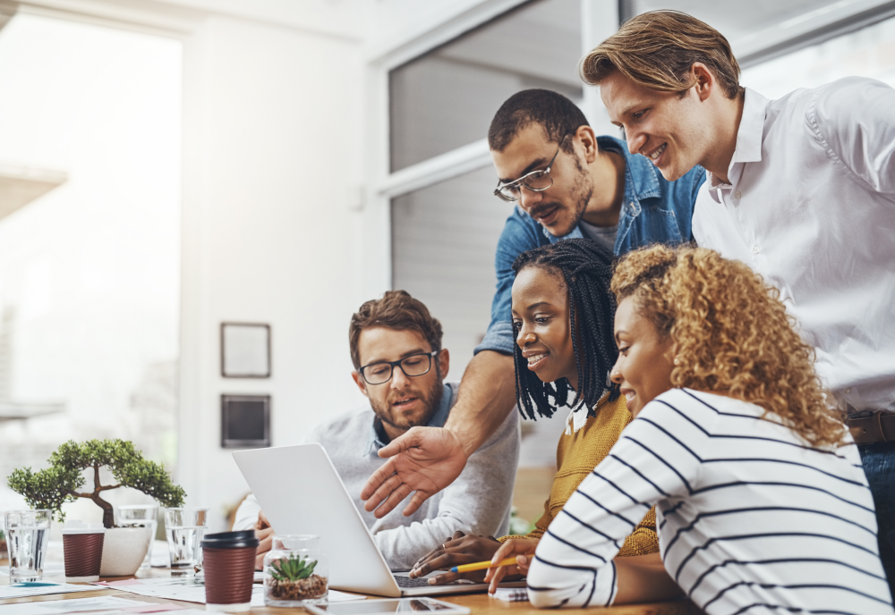 A group of co workers stand around a person on a laptop