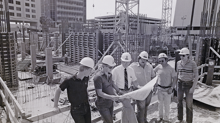 Six men of varying ages stand atop a building, wearing hard hats and viewing architectural plans together.