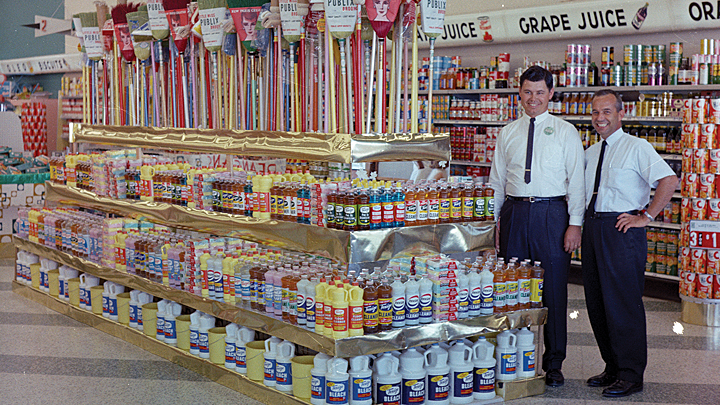 Two young men in black pants an white shirts with skinny ties stand by a colorful display of cleaning products and brooms in a Publix supermarket.