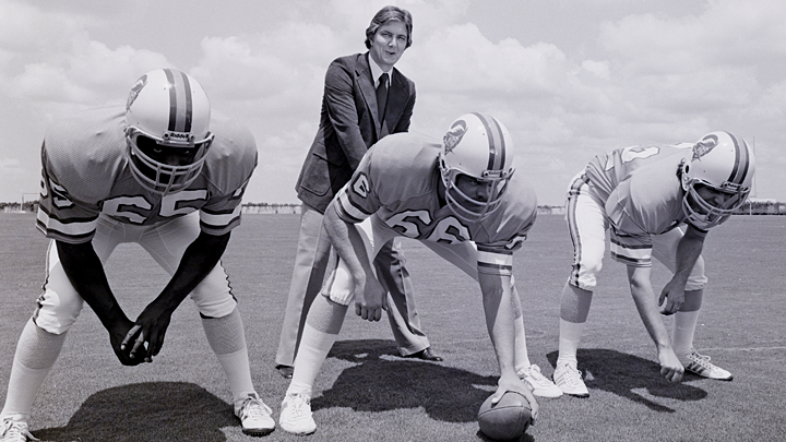 A black and white view of a tall young man in a suit jacket and light pants, sporting a 70s shag haircut, standing behind three Tampa Bay bucs players in uniform, who are about to hike the ball to him.