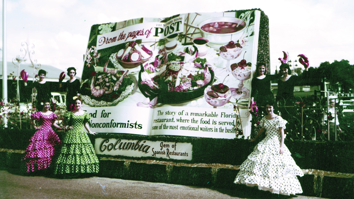 Three young women wearing ruffled floor-length dresses with puffed sleeves stand in front of a 10 ft. tall float designed like a book. The text says “From the pages of the Post” on a page with shrimp salad, soup and parfaits.
