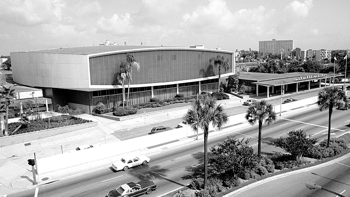 An aerial black and white view of a large, oblong, 60s-style public building with cars and palm trees in the foreground.