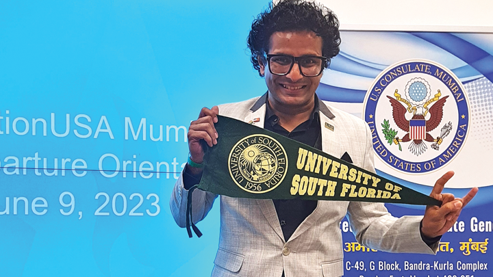 Ojas Rawal, a young Indian man with dark hair and glasses, stands in front of a bright blue display with Education USA Orientation in text. He is wearing a light gray suit and holding a University of South Florida pennant.