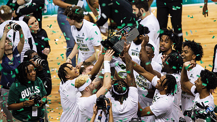 Men’s basketball players wearing USF uniforms and media stand amid a shower of green confetti on the court. They hold a large black and silver trophy over their heads.