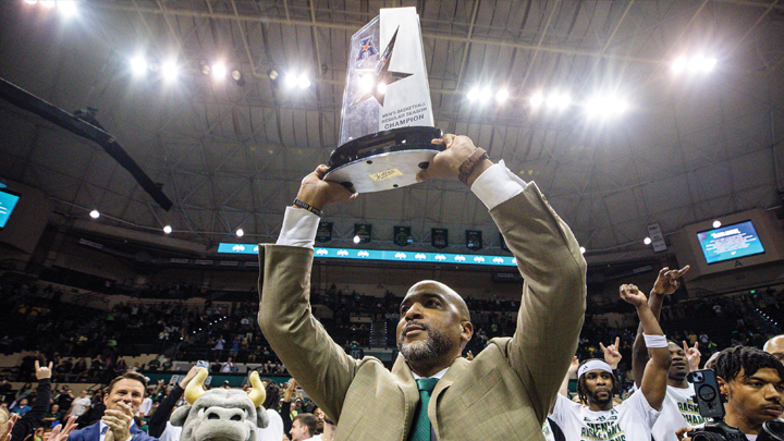 The coach, wearing a golden-tan jacket and deep USF green tie holds a large trophy over his head. The team, Rocky and Trustee Will Weatherford applaud and cheer in the background. 