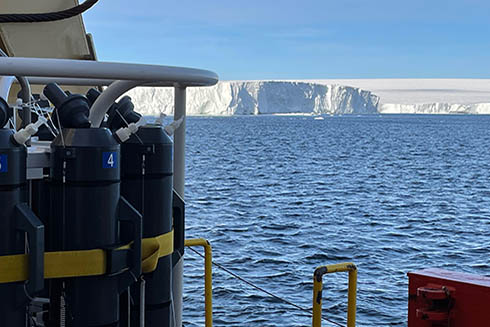 Sampling rosette with gray sampling bottles at left, the ship’s rail at lower right, and the face of the ice shelf in the background. Credit: Rob Sherrell