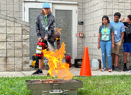 a student aims a fire extinguisher at a contained flame