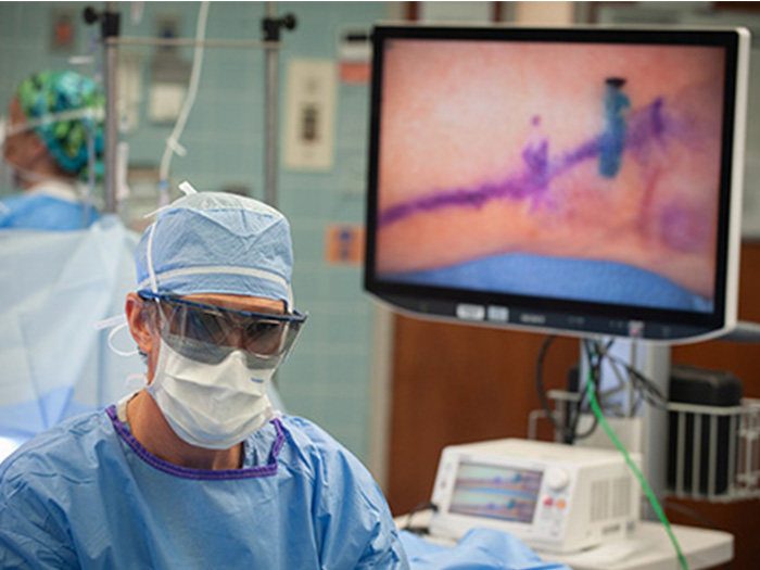 A surgeon wearing blue scrubs, a white face mask, and protective eye gear uses robotics during microsurgery.