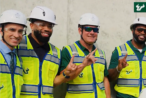 Four people in USF branded construction vests smile at the camera with the "Bull U" hand sign