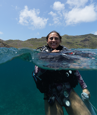 Tatiana in the ocean in scuba gear, smiling at camera which is half in water and half above