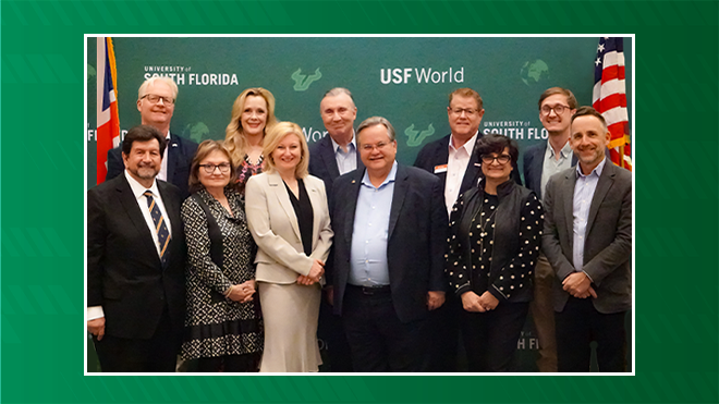 a group of usf and babc leadership smiling and standing in front of the green USF World backdrop with a british flag on the left and american flag on right