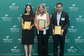 two females and a male dressed in professional wear pose infront of a USF backdrop while displaying their Faculty Teaching Award plaques